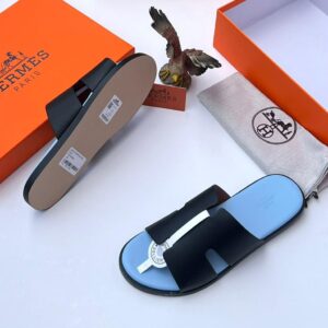 High-quality image of black leather Hermes Paris slide sandals with blue insole, displayed with orange box, dust bag, and eagle figurine on a white background.