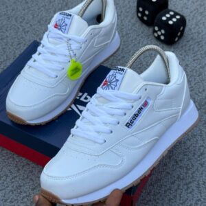 A pair of pure white Reebok Classic Leather Sneakers displayed on a grey surface with a shoe box and dice in the background, highlighting the shoe's sleek design and comfortable features.