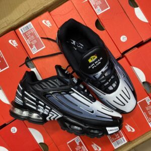 A pair of black and white Nike Air Max Plus sneakers on top of red shoe boxes, showcasing the shoe's sleek design and innovative technology.