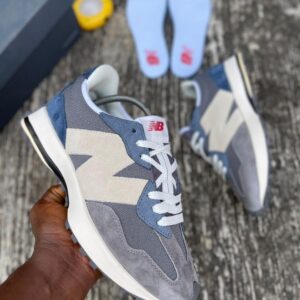 A pair of grey, white, and blue New Balance 574 sneakers, with a hand holding one shoe and the other on the ground, showcasing the shoe's sleek design and vibrant colors.