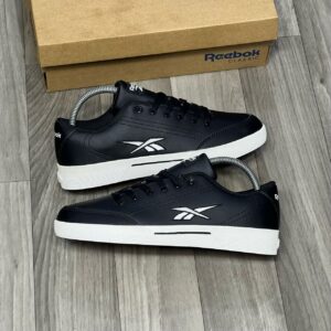 A pair of black Reebok Classic Leather Sneakers with white soles and logo, placed on a wooden floor next to a brown shoe box.