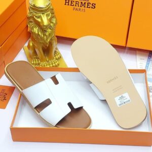 A pair of black leather slide sandals with Hermes Paris branding, nestled in an orange box with a golden lion statue in the background, evoking a sense of luxury and refinement.