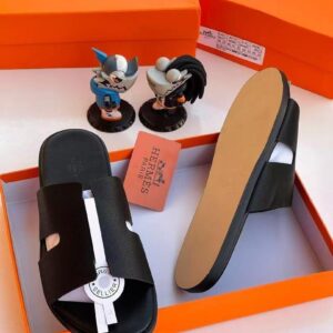 A pair of black leather slide sandals with a silver accent, displayed in an orange Hermes box with a dust bag and two figurines on a white background.