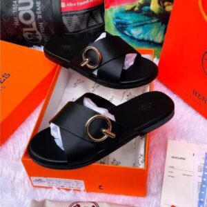 A pair of black leather slide sandals with a gold buckle, nestled in an orange box with Hermes branding, surrounded by luxury shopping bags.