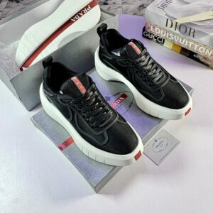 A pair of black Prada sneakers with red accents and white soles, displayed on shoe boxes with luxury brand books in the background.