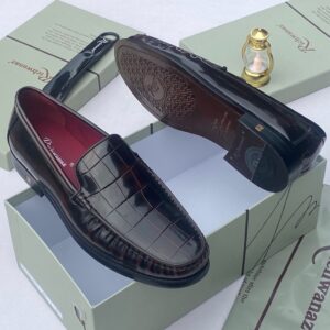A pair of black crocodile pattern loafers with red interior, displayed on a shoe box with a luxurious background.