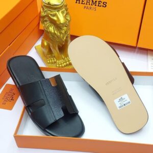 A pair of black leather slide sandals with Hermes Paris branding, nestled in an orange box with a golden lion statue in the background, evoking a sense of luxury and refinement.