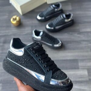 Close-up of a hand holding a black sneaker with rhinestones and silver accents, with two more pairs in the background.