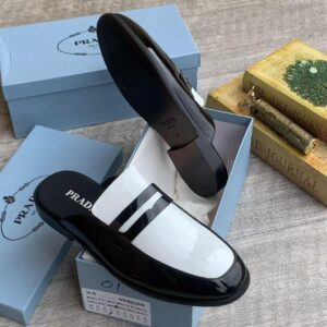 A pair of black patent leather loafers with white insoles and Prada branding, displayed in a light blue shoe box with a luxurious setting.