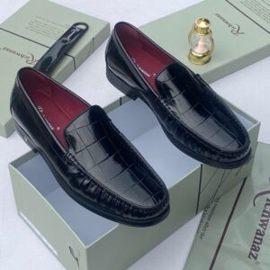 A pair of black crocodile pattern loafers with red interior, displayed on a shoe box with a luxurious background.