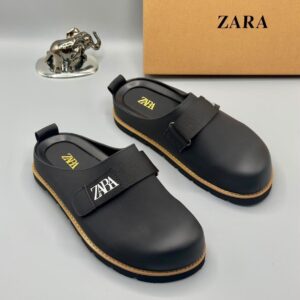 A pair of luxury black leather clogs with golden accents and ZARA logo, displayed on a pristine white surface with a brown ZARA box and golden elephant figurine in the background, showcasing high-class products for the elite at Levonteluxe in Nigeria.