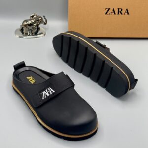A pair of luxury black leather clogs with golden accents and ZARA logo, displayed on a pristine white surface with a brown ZARA box and golden elephant figurine in the background, showcasing high-class products for the elite at Levonteluxe in Nigeria.