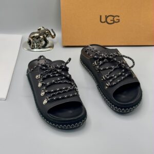 Luxury black UGG sandals with metal buckles and laces on a white surface, accompanied by a UGG box and a silver elephant statue, epitomizing high-class style and elegance.