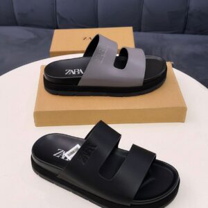 A pair of luxurious ZARA slides, one in black and the other in black and grey, displayed on a white table with a brown shoe box in the background.